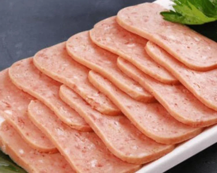 Slices of processed ham arranged on a white plate with a green garnish in the background.