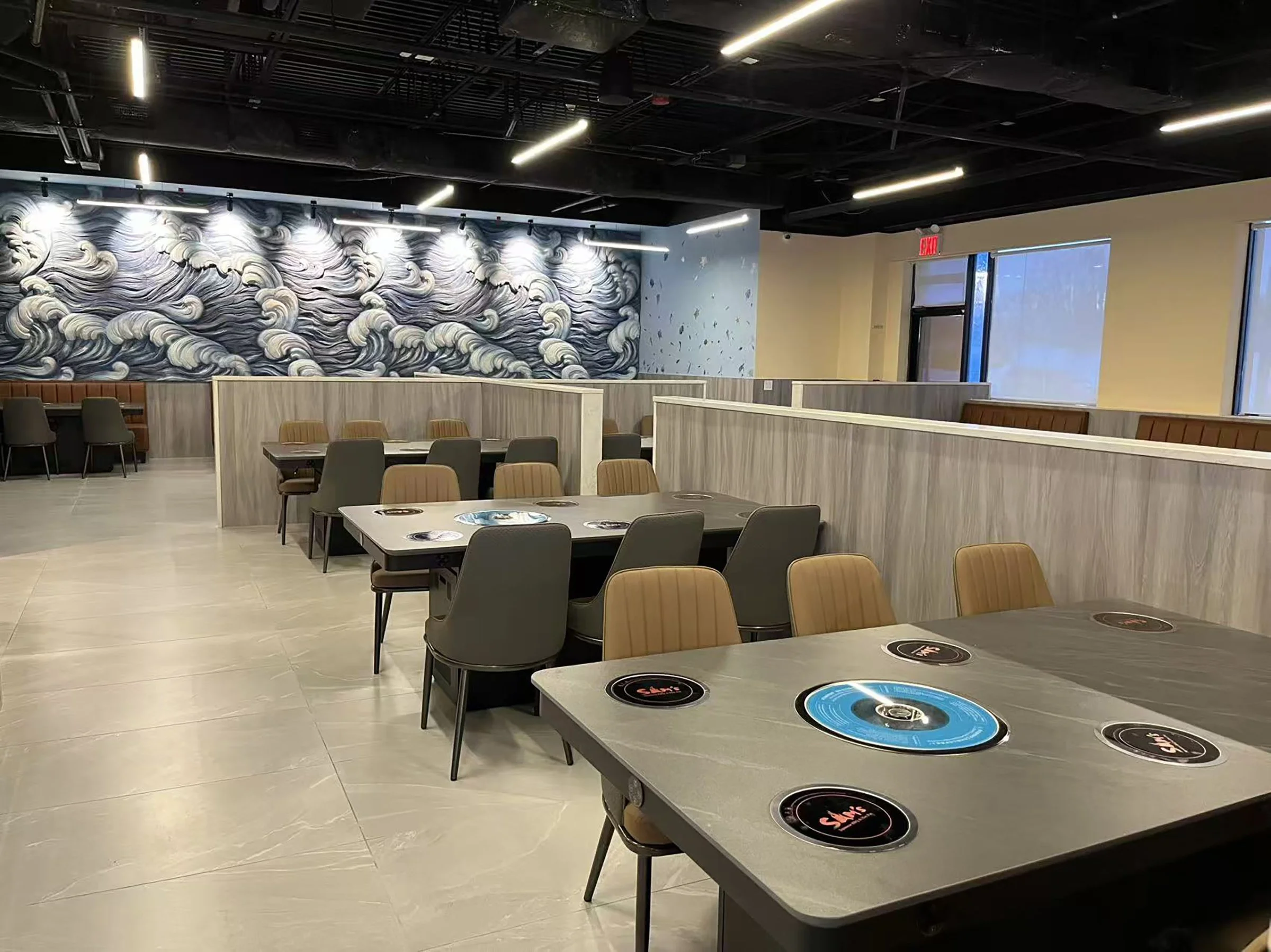 Empty restaurant with tables and chairs, large ocean wave mural on wall, modern lighting, and private booths.