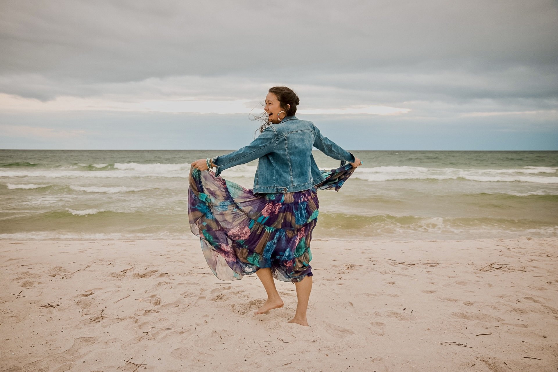 Woman in a denim jacket and colorful flowing dress dancing on a sandy beach with ocean waves and cloudy sky in the background.