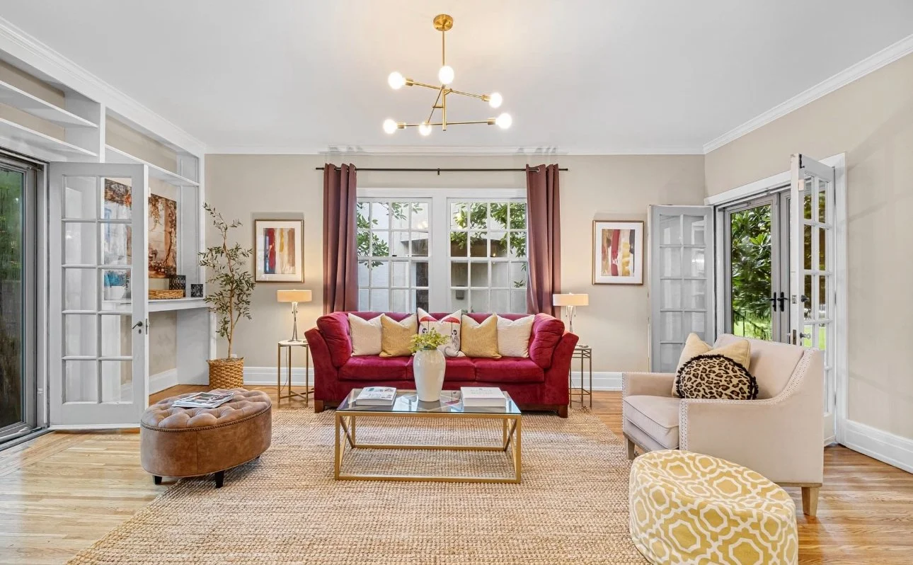Bright living room with a red sofa, beige armchair with leopard print pillow, glass-top coffee table, and large windows with pink curtains.