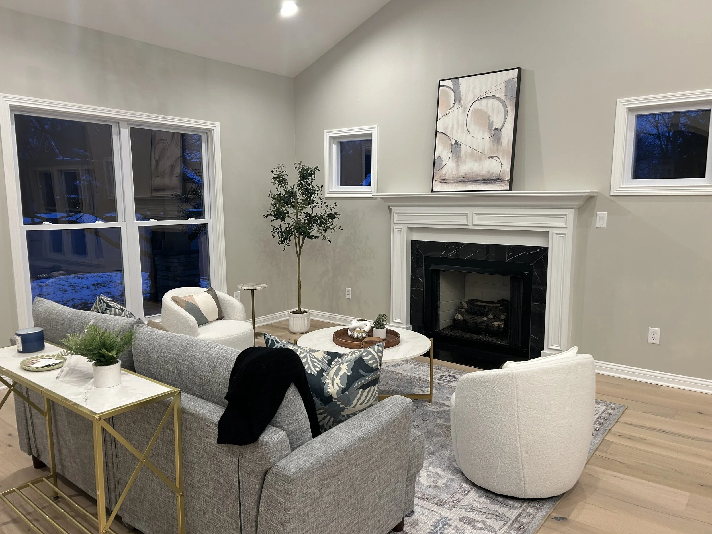 Living room with a white fireplace, abstract wall art, light-colored walls, and large windows showing a snowy outdoor scene. The room has a gray sofa with decorative pillows, a white armchair, a gold side table with a plant, and a marble coffee table