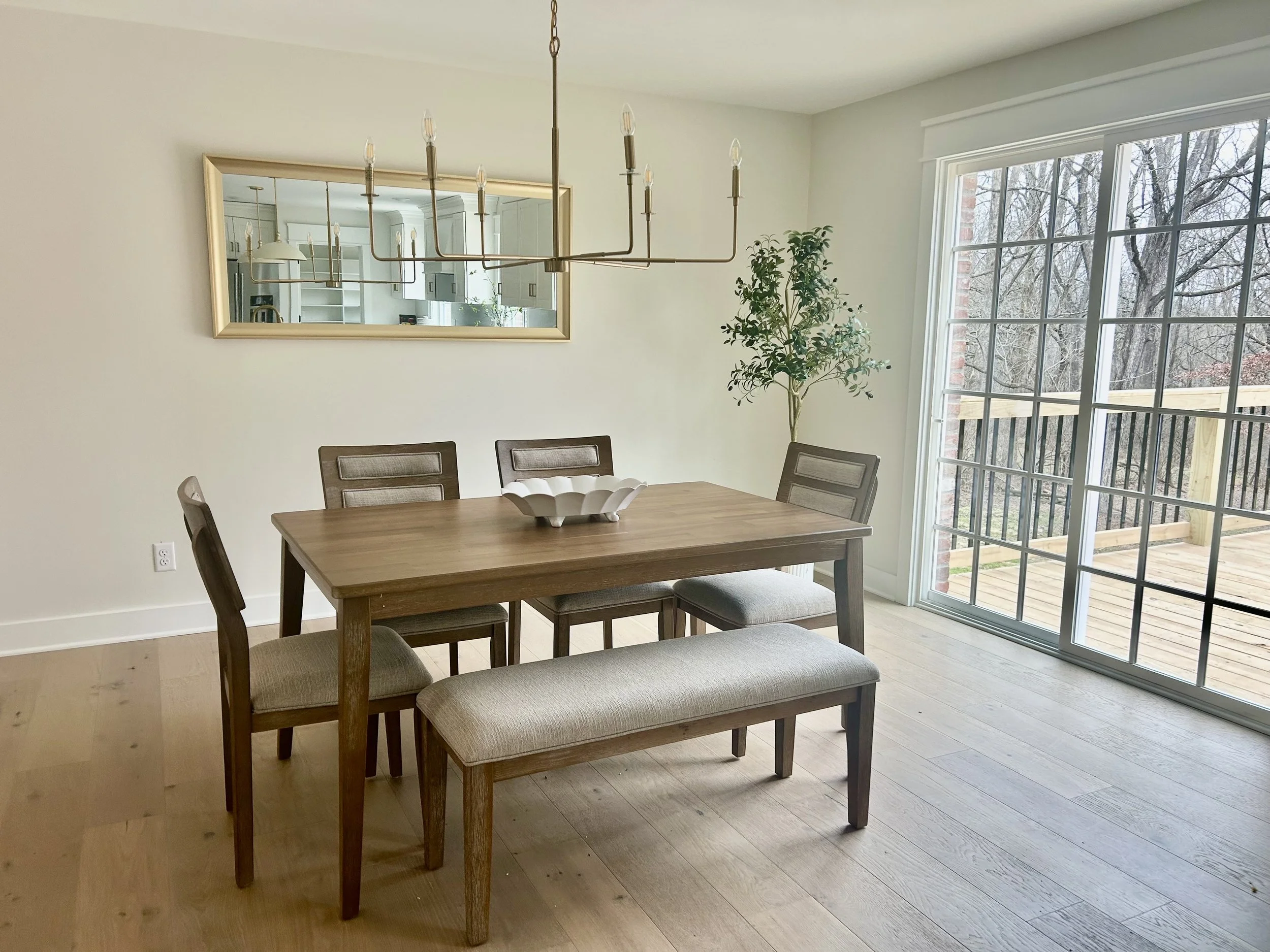 Dining room with a rectangular wooden table, four chairs, a window, a glass door leading to the deck, a large mirror on the wall, a chandelier hanging above, and a potted plant in the corner.
