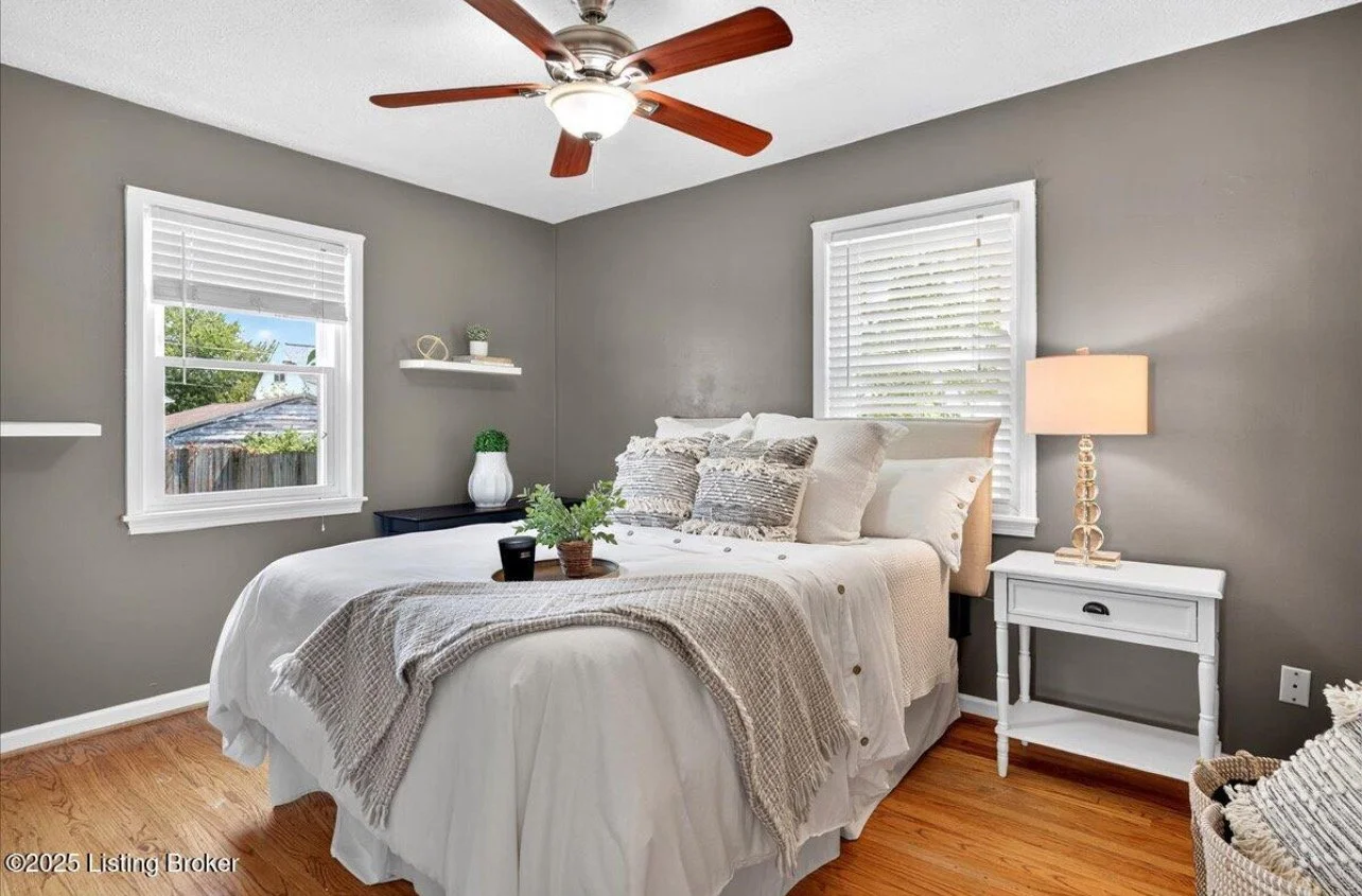 A cozy bedroom with a white bed, gray walls, wooden floors, a ceiling fan, two windows with white blinds, a white bedside table with a lamp, and decorative pillows.