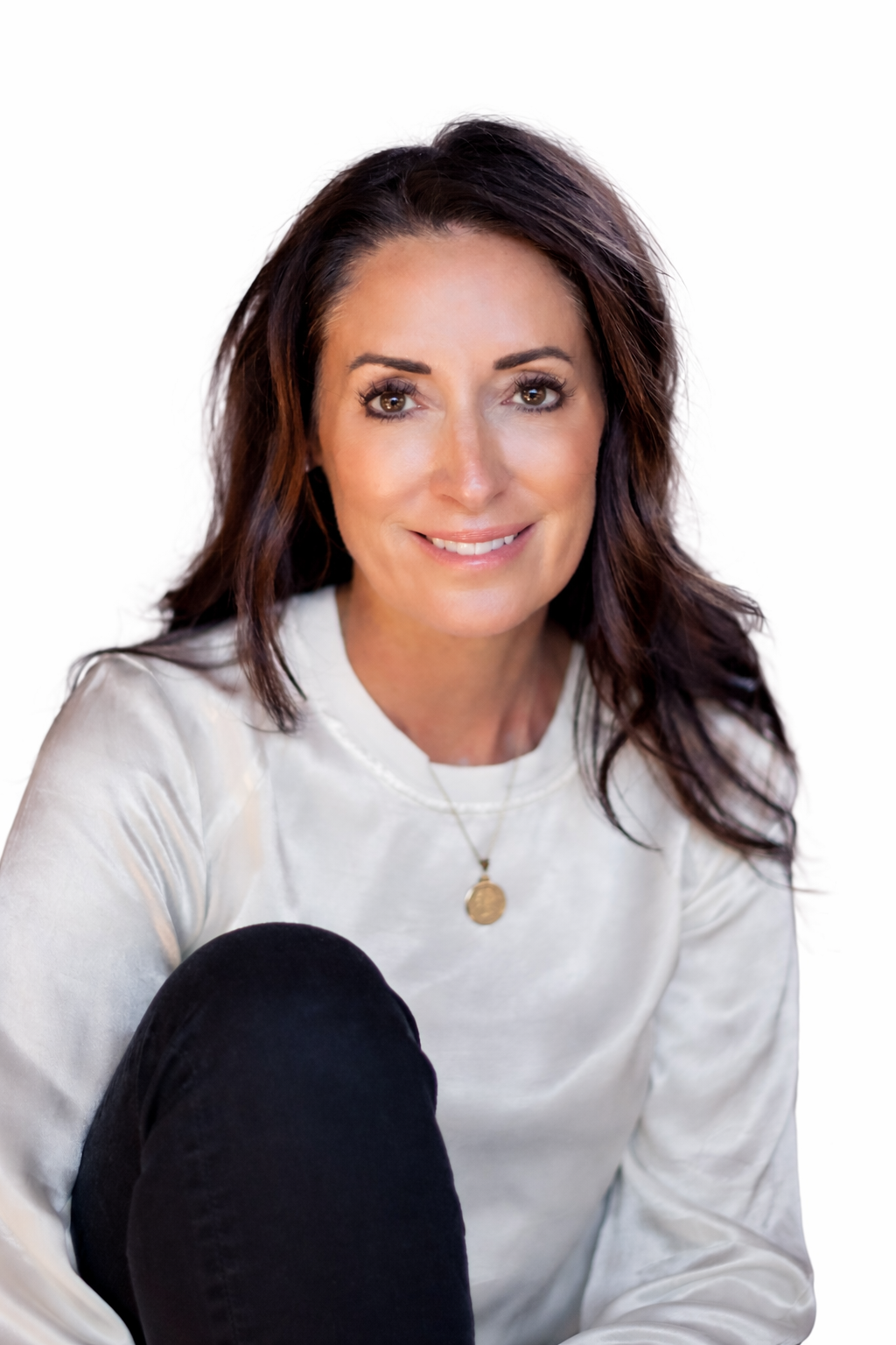 A woman with dark brown hair and brown eyes smiling, wearing a white long sleeve shirt and a gold pendant necklace, sitting with her knee raised, against a plain white background.