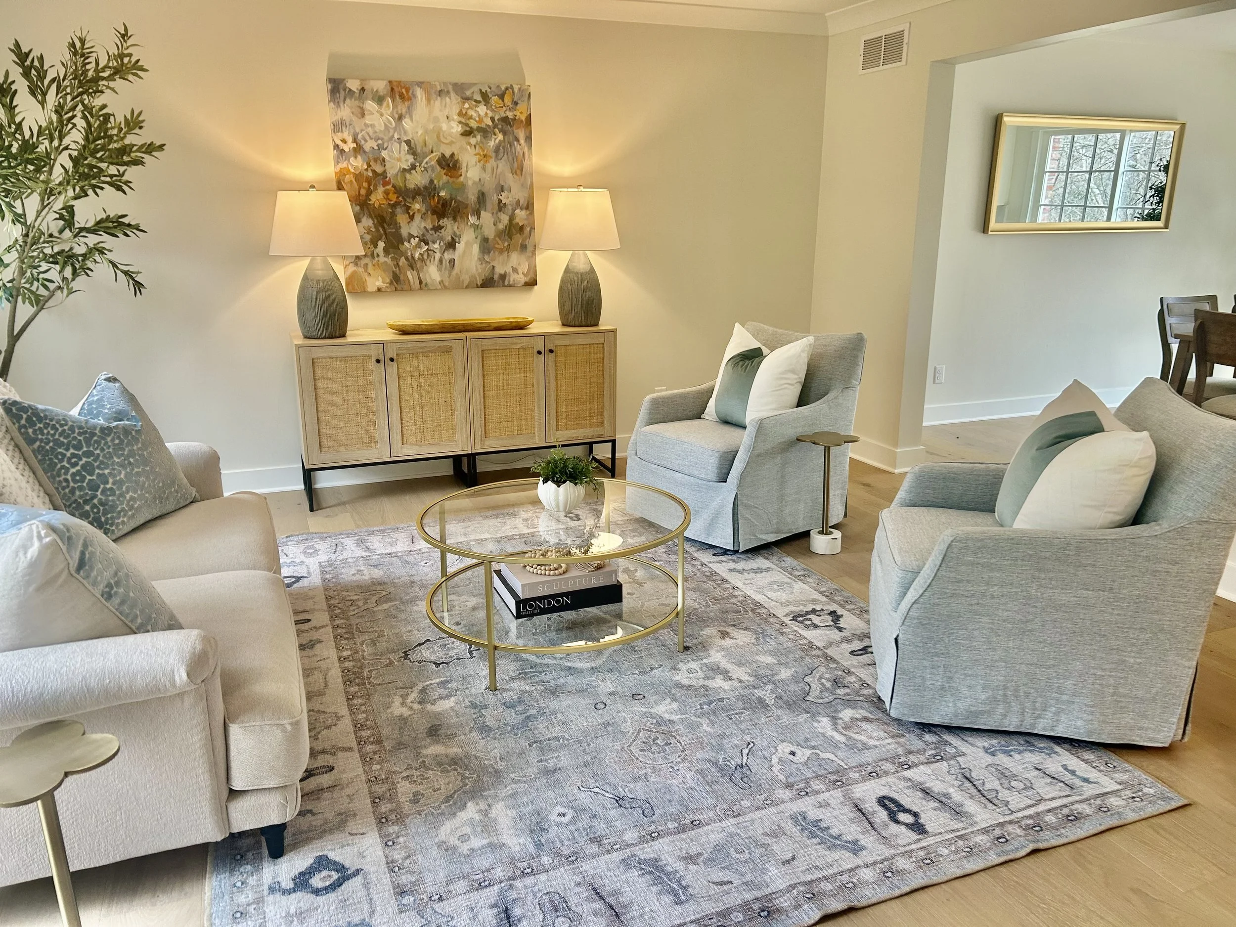 Living room with light-colored sofas and armchairs, a gold coffee table, a wooden sideboard with lamps, abstract wall art, a rug, and a mirror on the wall.