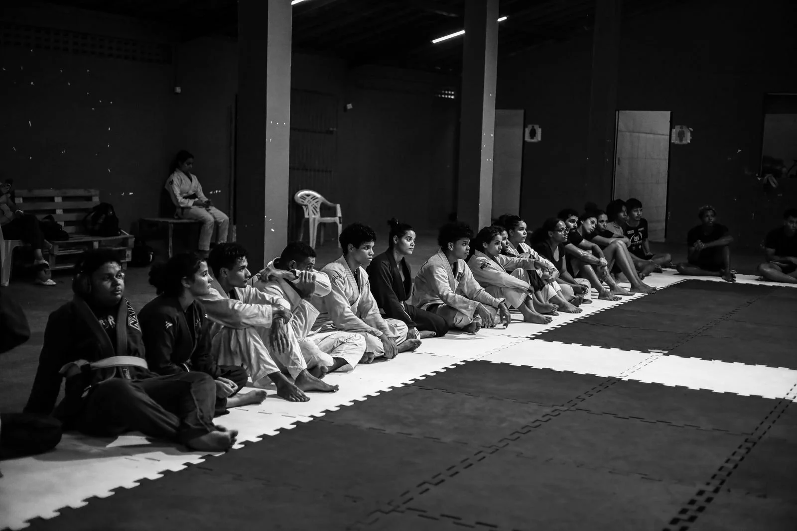 Grupo de pessoas sentadas em fila no tatame durante uma aula de jiu-jitsu, usando roupas tradicionais, em ambiente de ginásio com iluminação suave.