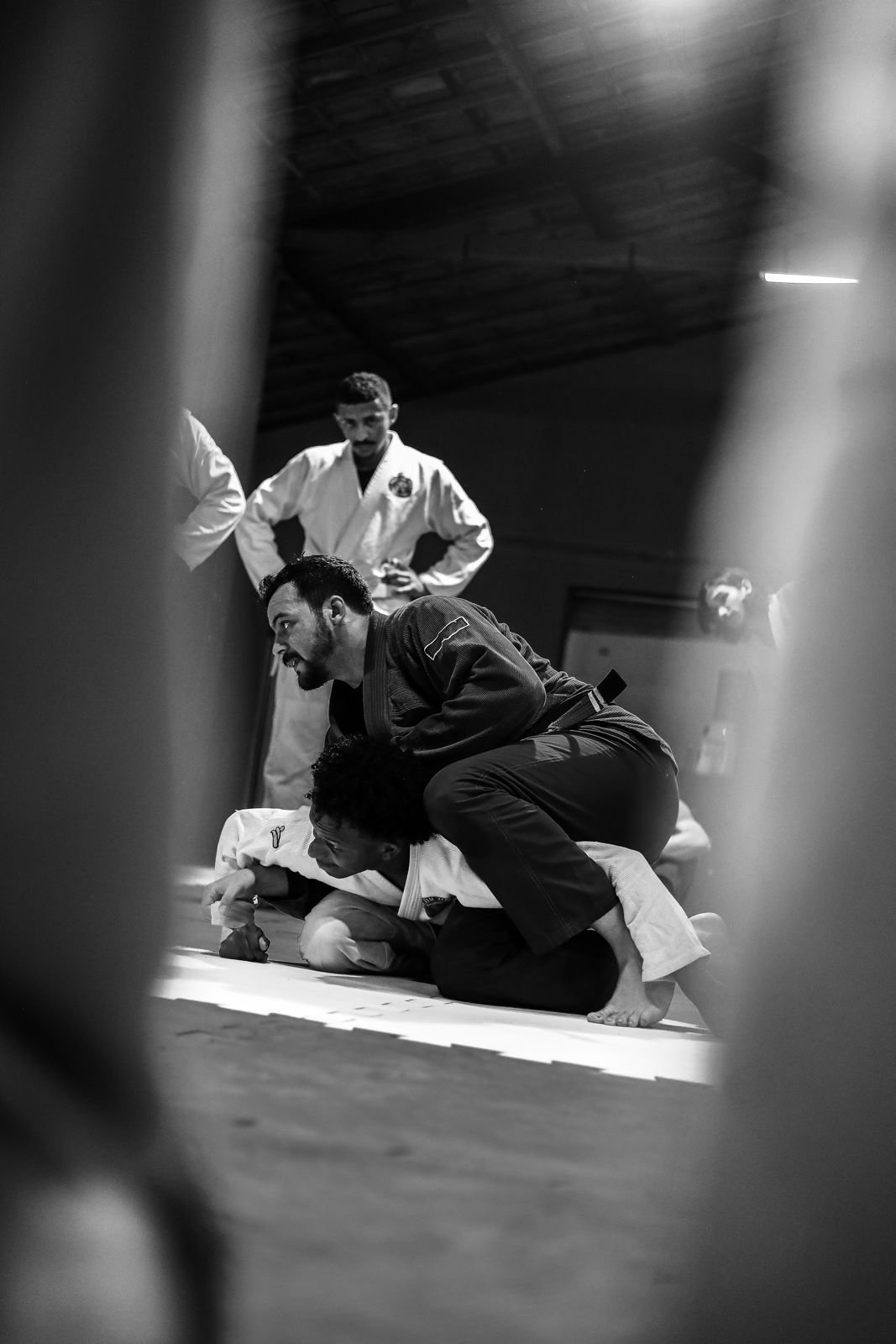 Duas pessoas treinando jiu-jitsu no tatame, com uma pessoa aplicando uma técnica em outra, enquanto outras observam ao fundo.