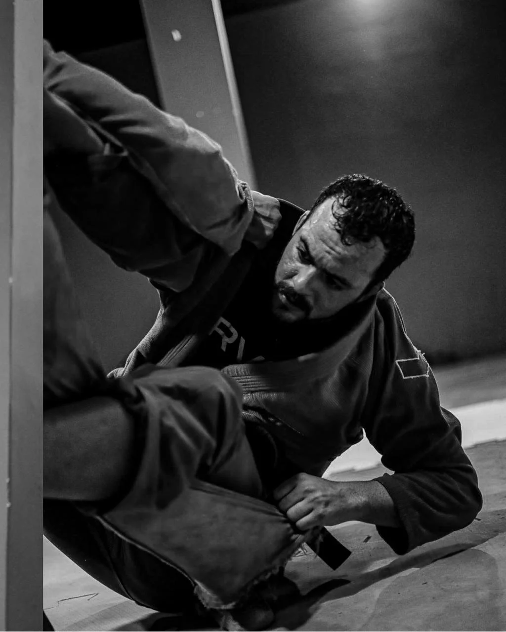 Judoca de kimono escuro no chão durante uma luta de jiu-jitsu, puxando a manga do oponente.
