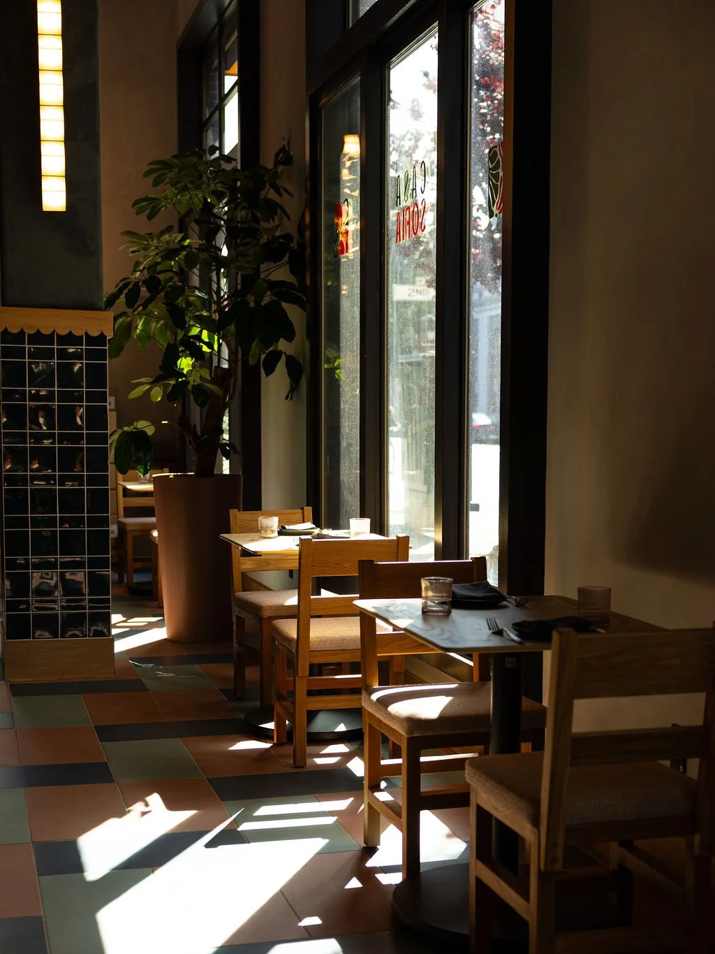 Empty restaurant with wooden tables and chairs, sunlight streaming through large windows, and a potted plant.