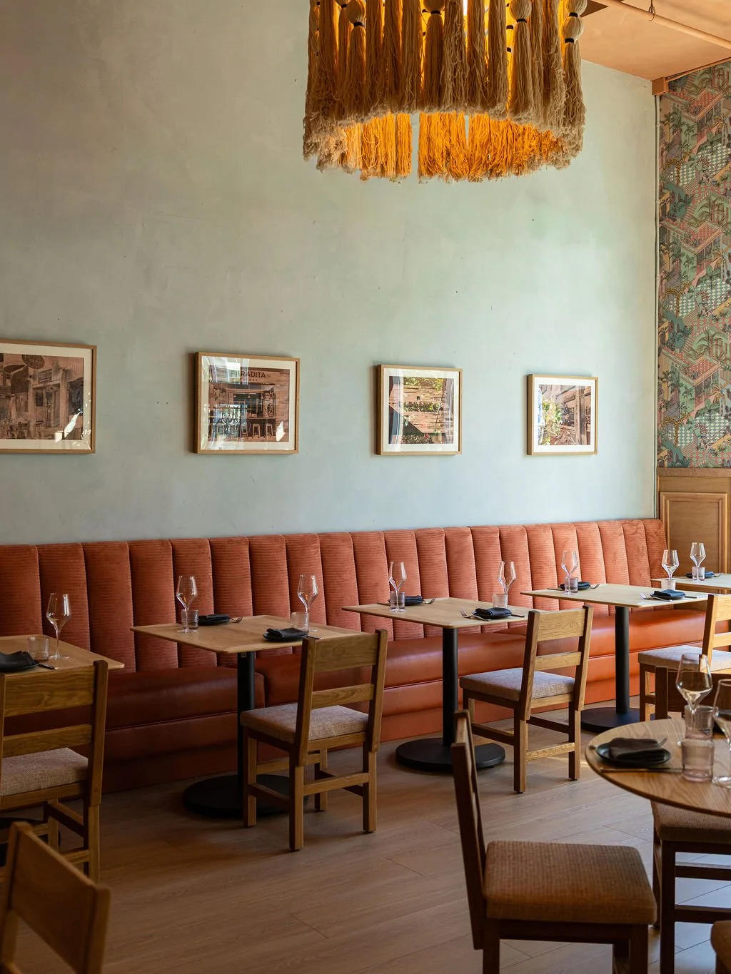 Empty restaurant dining area with a long rust-colored banquette, wooden tables, chairs, and framed pictures on the light green wall. The ceiling has a decorative yellow tassel chandelier.