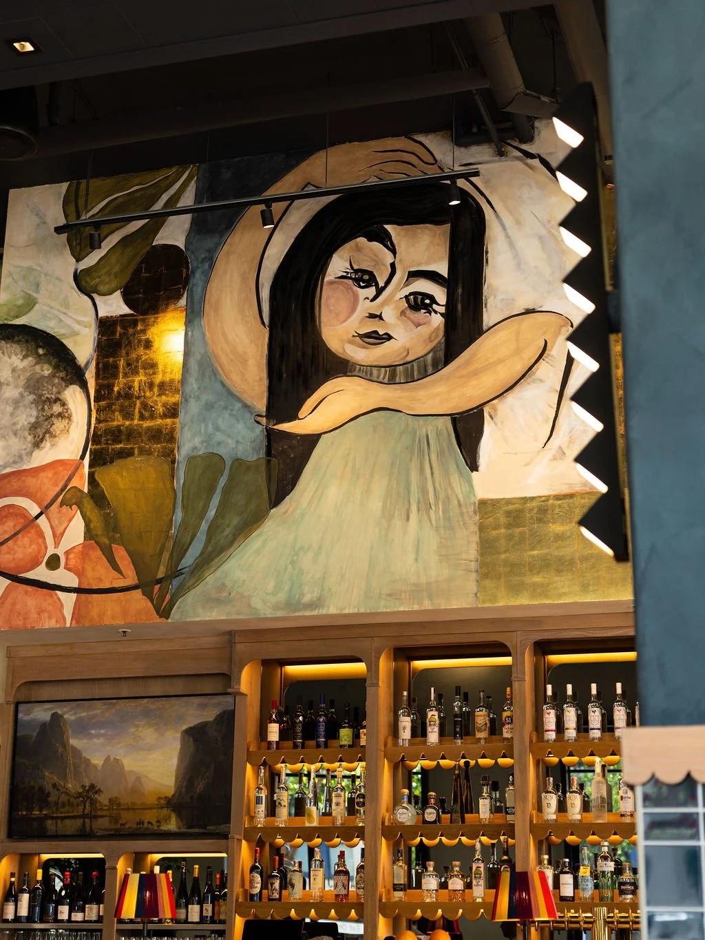 A colorful mural of a woman with long black hair, barefoot, wearing a beige dress, with her arms crossed and resting on her knees, is painted on a wall above a bar area with shelves of liquor bottles. The mural features warm and earthy tones with abs