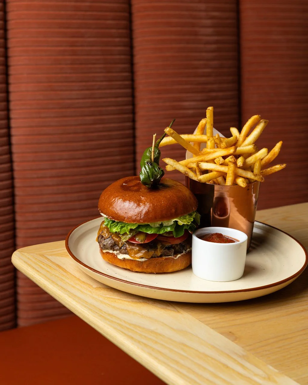 A burger with lettuce, tomato, cheese, and a beef patty on a bun, topped with a green chili on a plate. Next to it are French fries in a copper cup and a small white cup of ketchup on a wooden table.