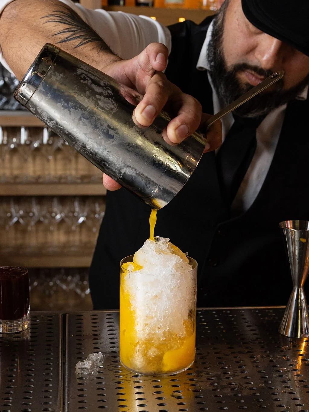 Bartender pouring a yellow cocktail over crushed ice into a glass at a bar.
