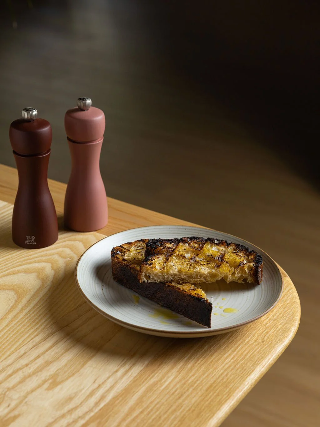 A slice of grilled bread with a topping, on a white plate, placed on a light wooden table, next to two salt and pepper shakers.