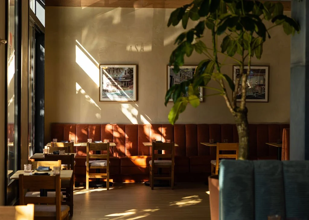 Interior of a restaurant with wooden tables and chairs, sunlight streaming through windows casting shadows, framed pictures on the wall, and a large potted tree in the foreground.