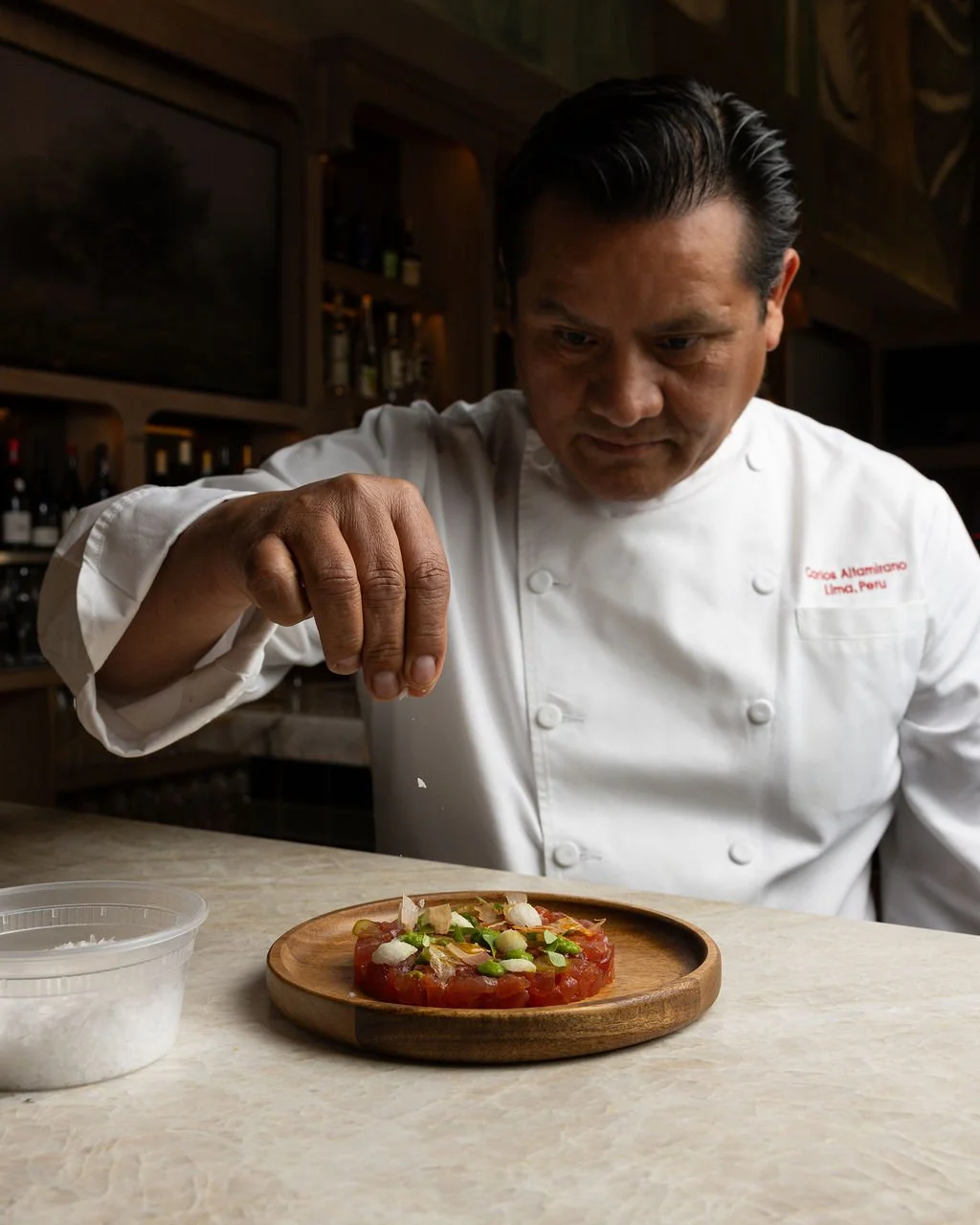Chef adding finishing touches to a dish, which appears to be a tartare, on a wooden plate in a restaurant kitchen.