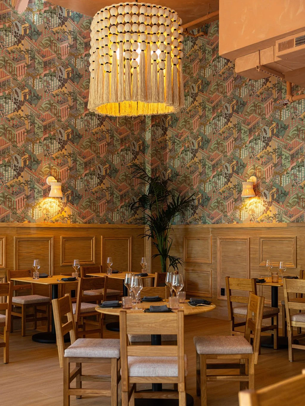 A cozy restaurant interior with wooden chairs, tables set with wine glasses, cloth napkins, and water glasses. The room features a decorative patterned wallpaper, wall-mounted lights, a large fringed chandelier, and a tall plant in the corner.