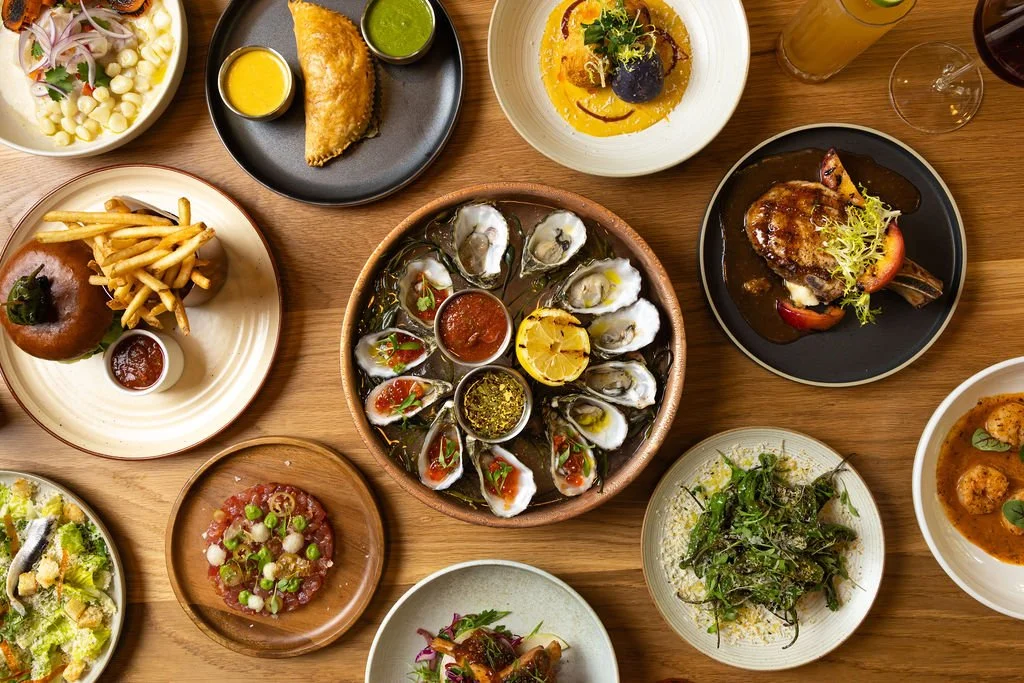 A variety of dishes including oysters, a salad, fries, a steak served with vegetables, and several appetizers on a wooden table.