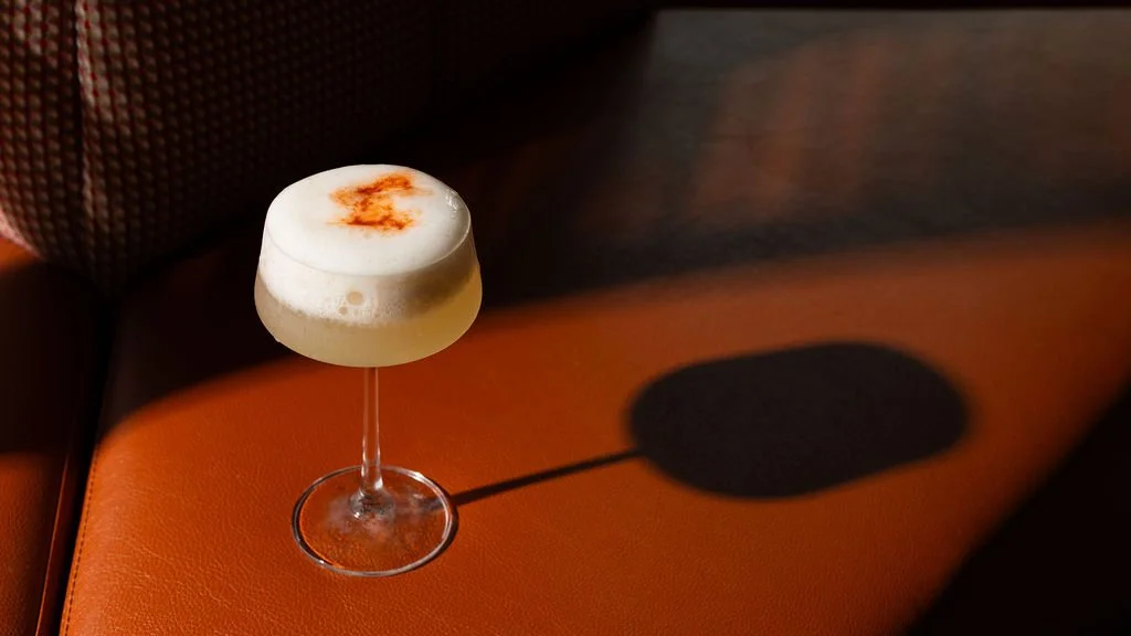 A cocktail glass with a creamy frothy drink topped with a sprinkle of reddish-brown spice, placed on a leather surface with a shadow cast beside it.