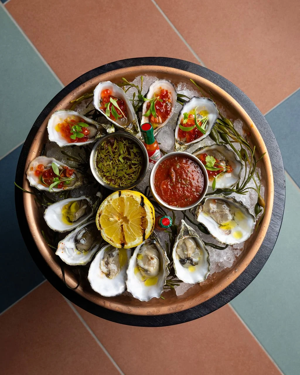 A round tray of fresh oysters on ice, garnished with herbs and lemon slices, with small bowls of cocktail sauce and pickled vegetables in the center, on a tiled floor.