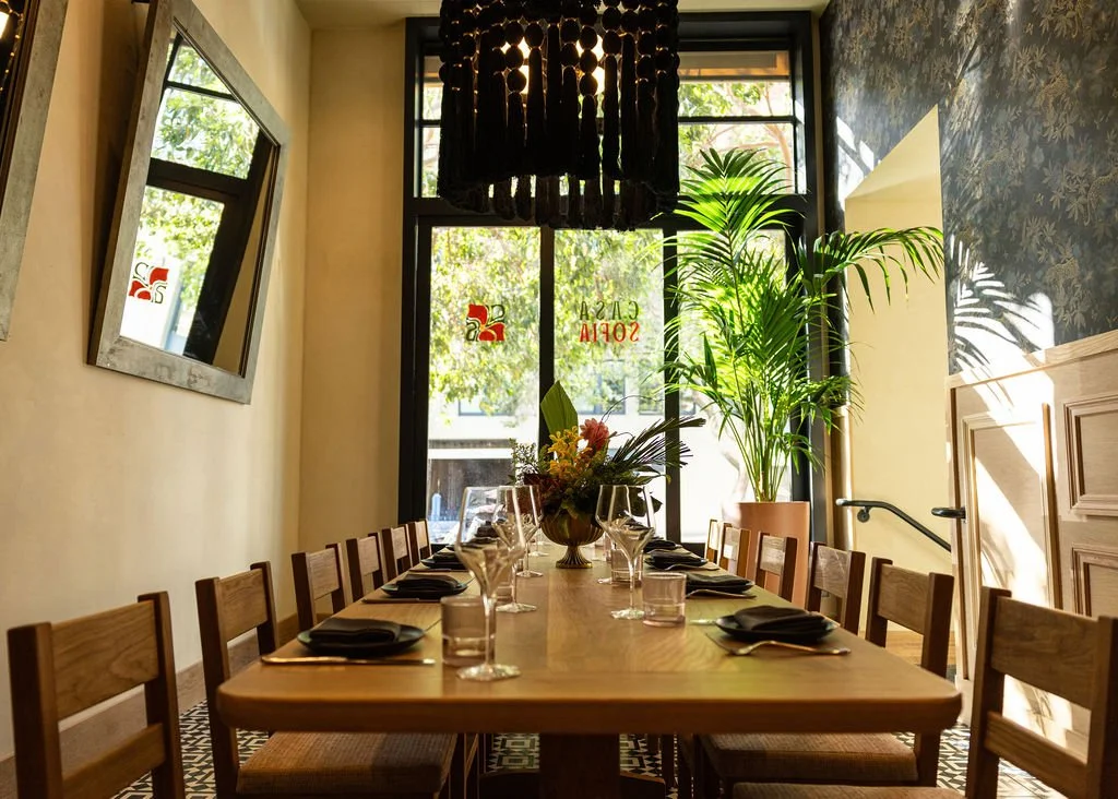 A long wooden dining table set with black napkins, wine glasses, water glasses, and a floral centerpiece, in a bright room with large windows, potted plants, and decorative wall mirrors.