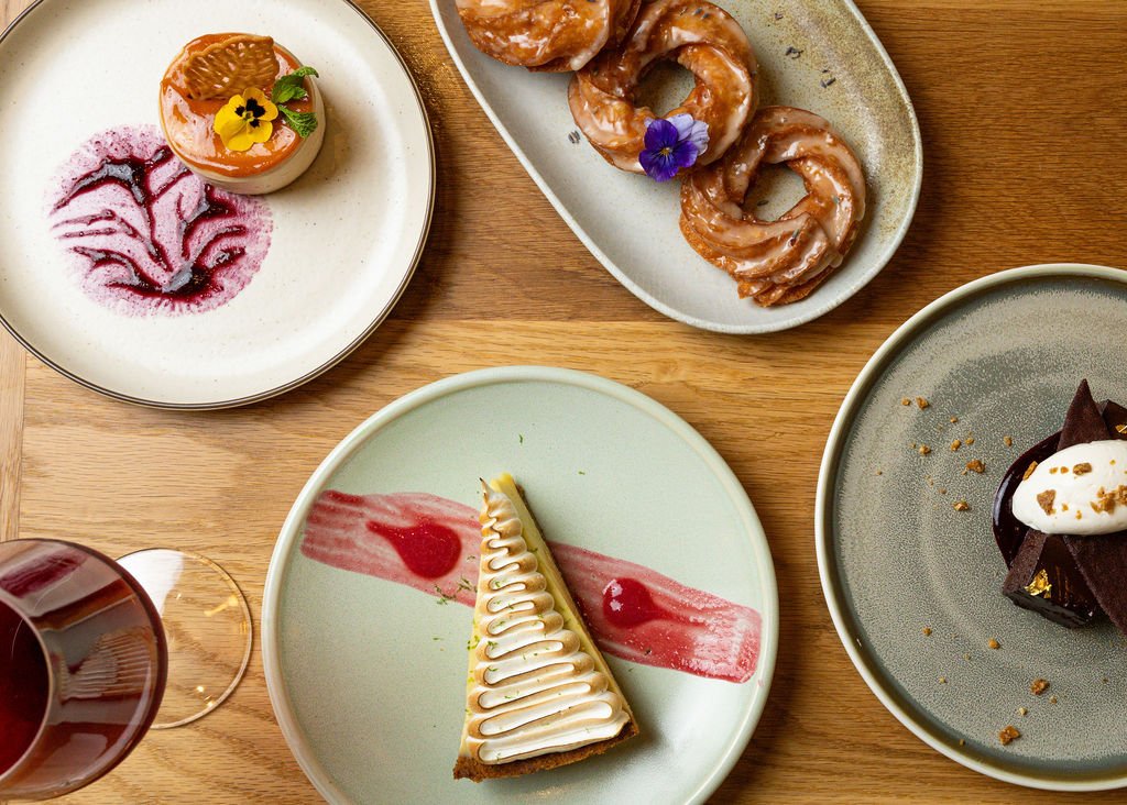 Assorted desserts and a glass of red wine on a wooden table, including a cheesecake with a red sauce, a chocolate cake with cream, a slice of lemon meringue pie, and caramelized donuts topped with edible flowers.