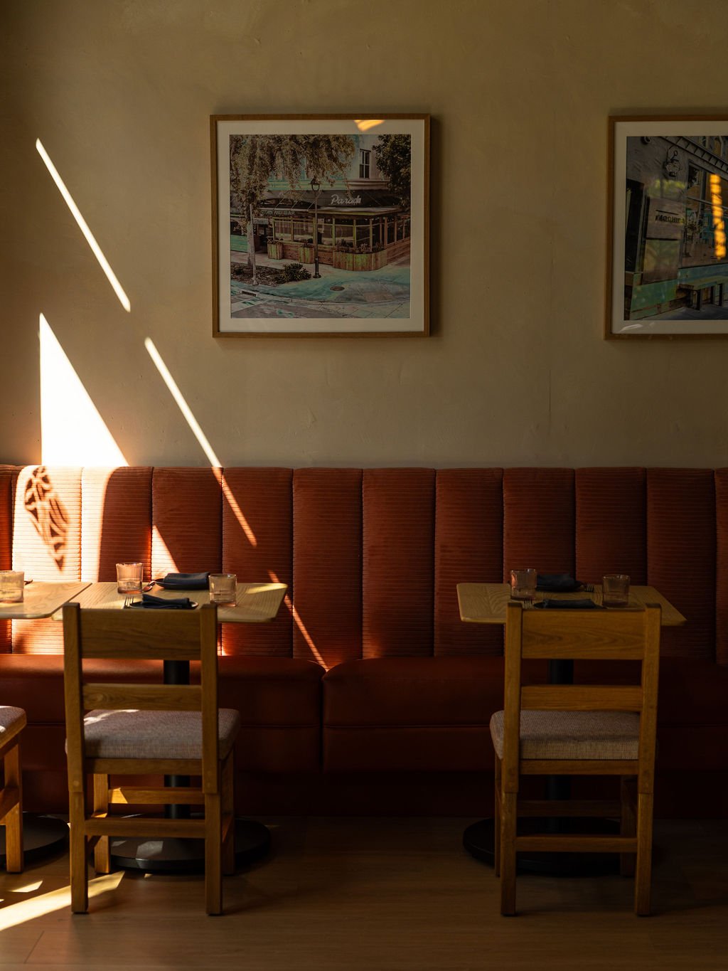 Sunlight casting shadows on a restaurant wall, three framed pictures hanging above a red upholstered bench, wooden tables set with napkins and glasses, and wooden chairs at the tables.
