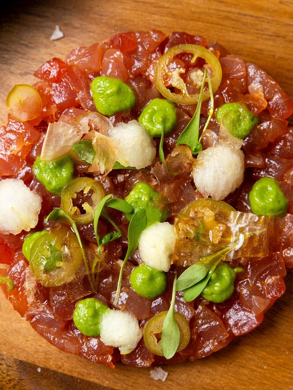 A close-up of a steak tartare garnished with green peas, sliced green chili peppers, microgreens, pickled onions, and thin flakes of cheese on a wooden surface.