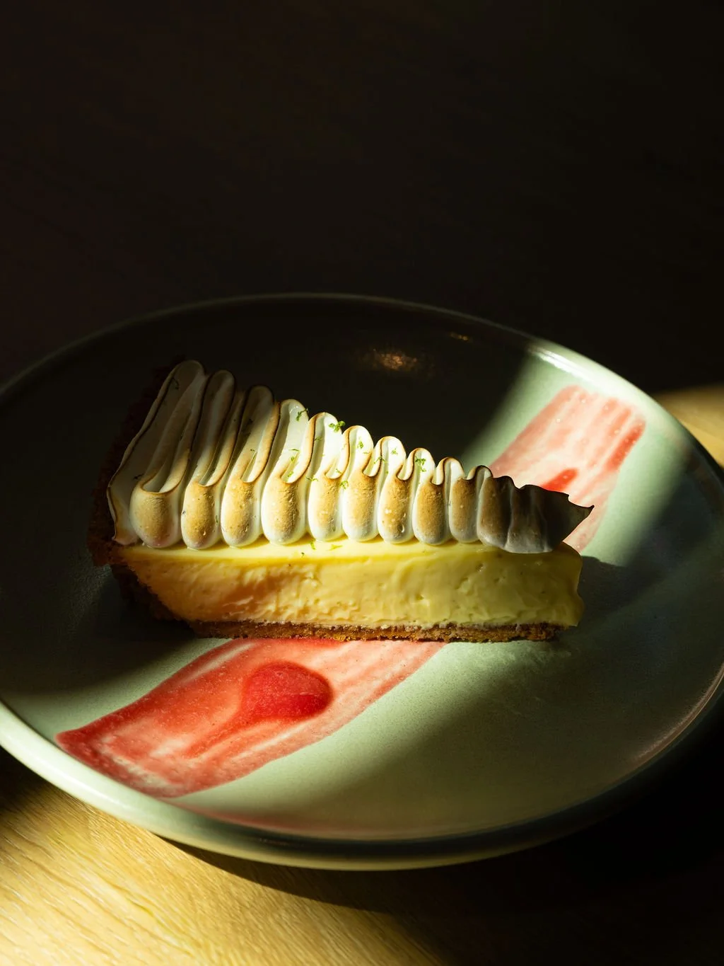 A slice of lemon meringue pie on a decorative plate with pink and red watercolor accents, placed on a wooden surface, with sunlight highlighting the dessert.