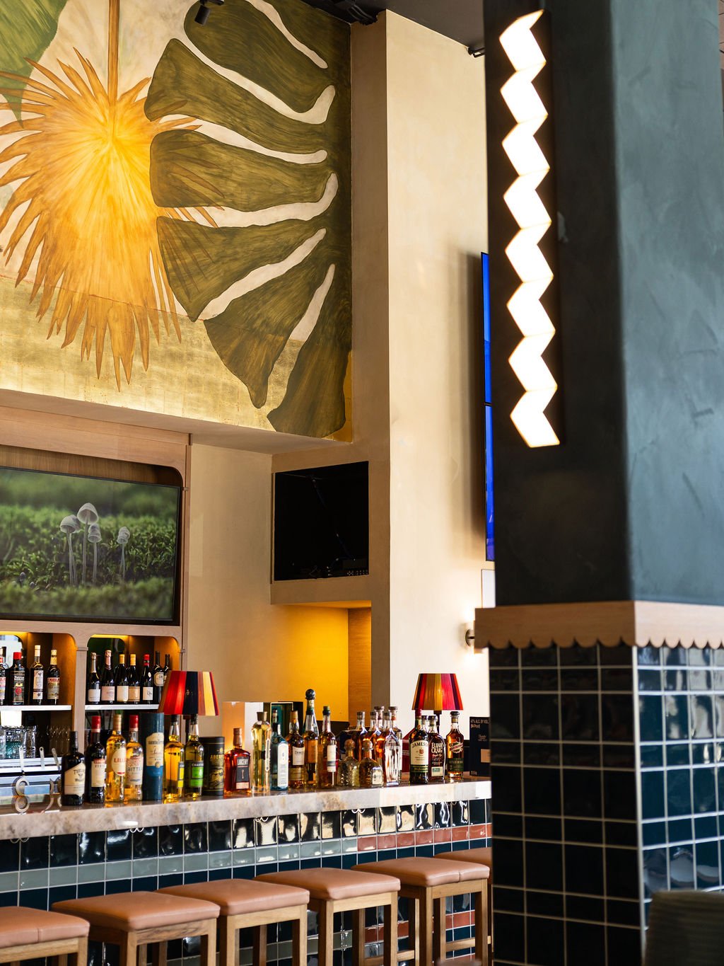 Bar area with liquor bottles, bar stools, a large leafy mural on the wall, and a television screen showing mushrooms.