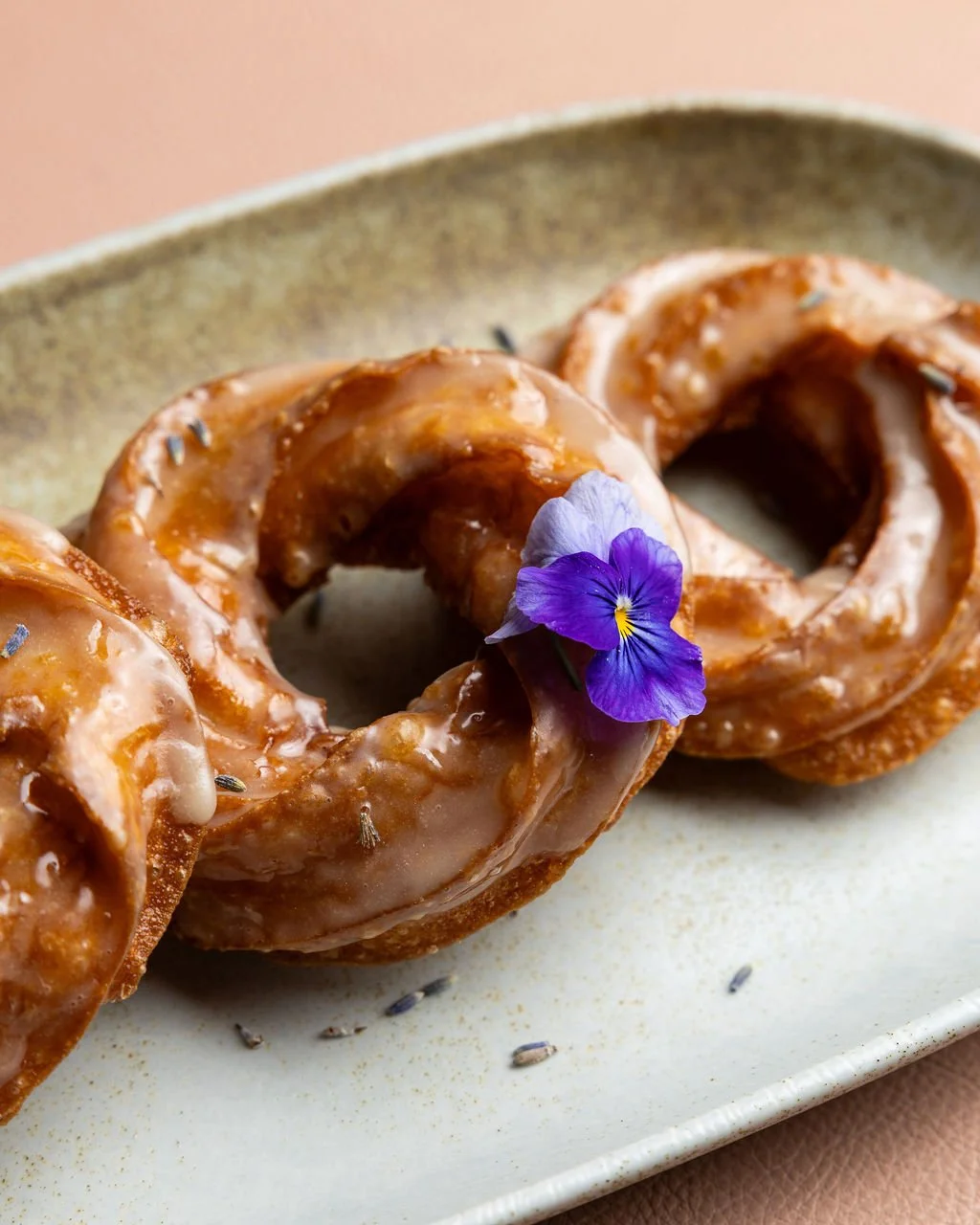 Glazed donuts garnished with a purple edible flower on a ceramic plate.