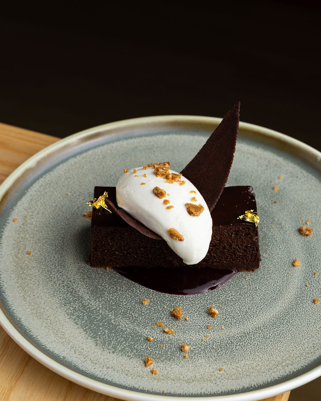 Chocolate cake with a dark chocolate triangle, whipped cream, and crushed nuts, served on a gray ceramic plate.