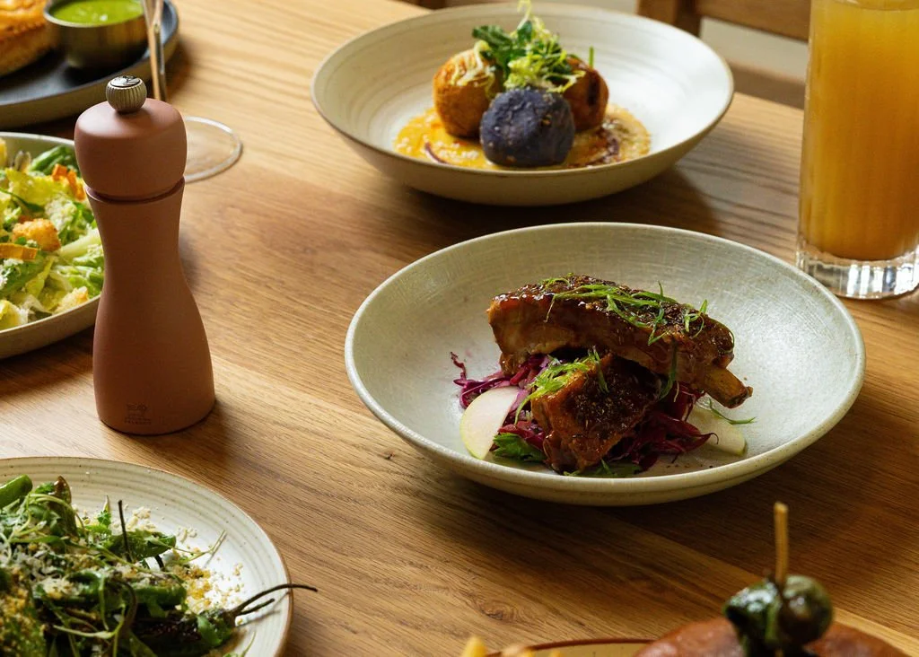 Assorted dishes on a wooden table, including salad, lamb chops with vegetables, and a plate with croquettes and sauce, along with a glass of beer and a salt grinder.