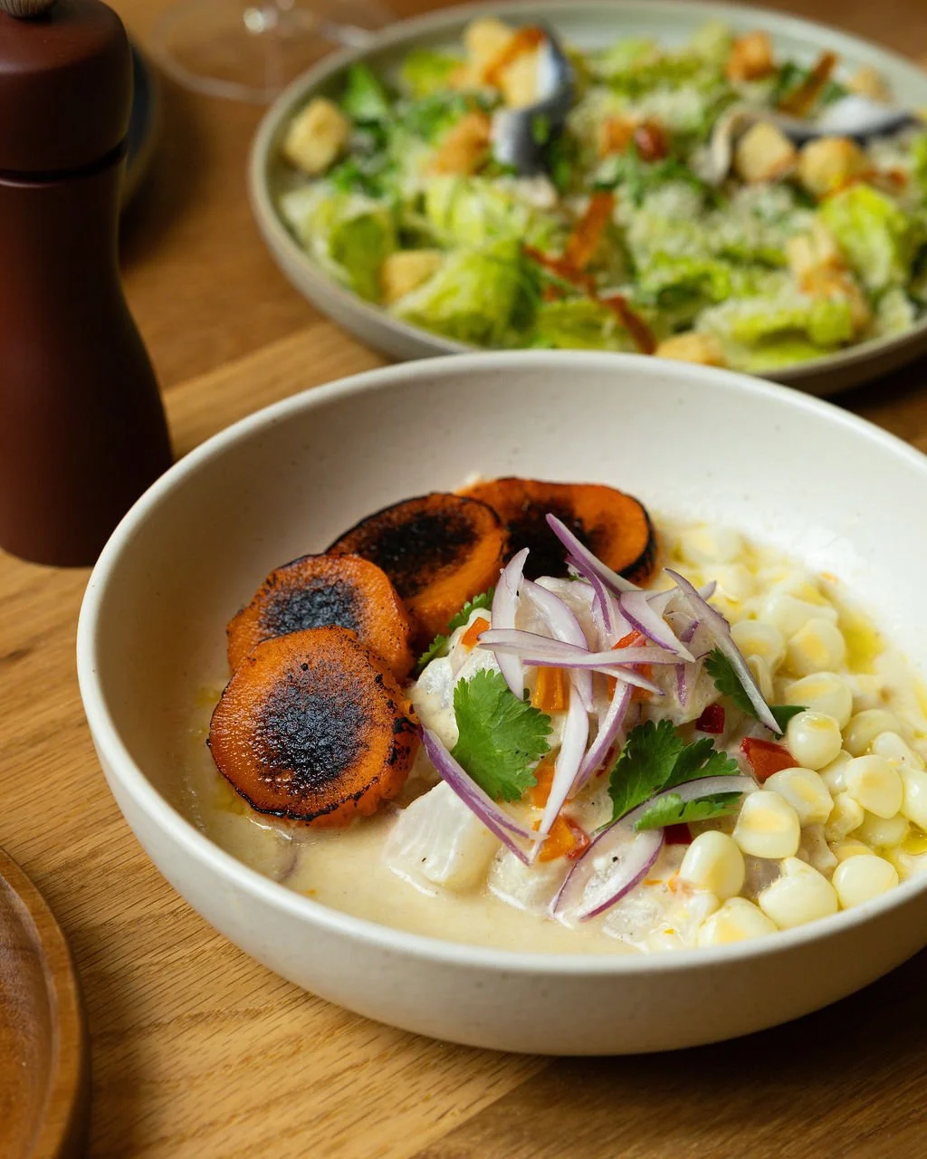 Bowl of grilled plantains, corn, and chopped vegetables, garnished with cilantro and sliced red onions, on a wooden table. In the background, a green salad with croutons and shredded cheese in a bowl.