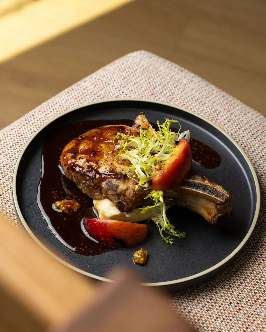 A plate with a cooked lamb chop topped with greens and apple slices, served with sauce and mustard on a black dish.