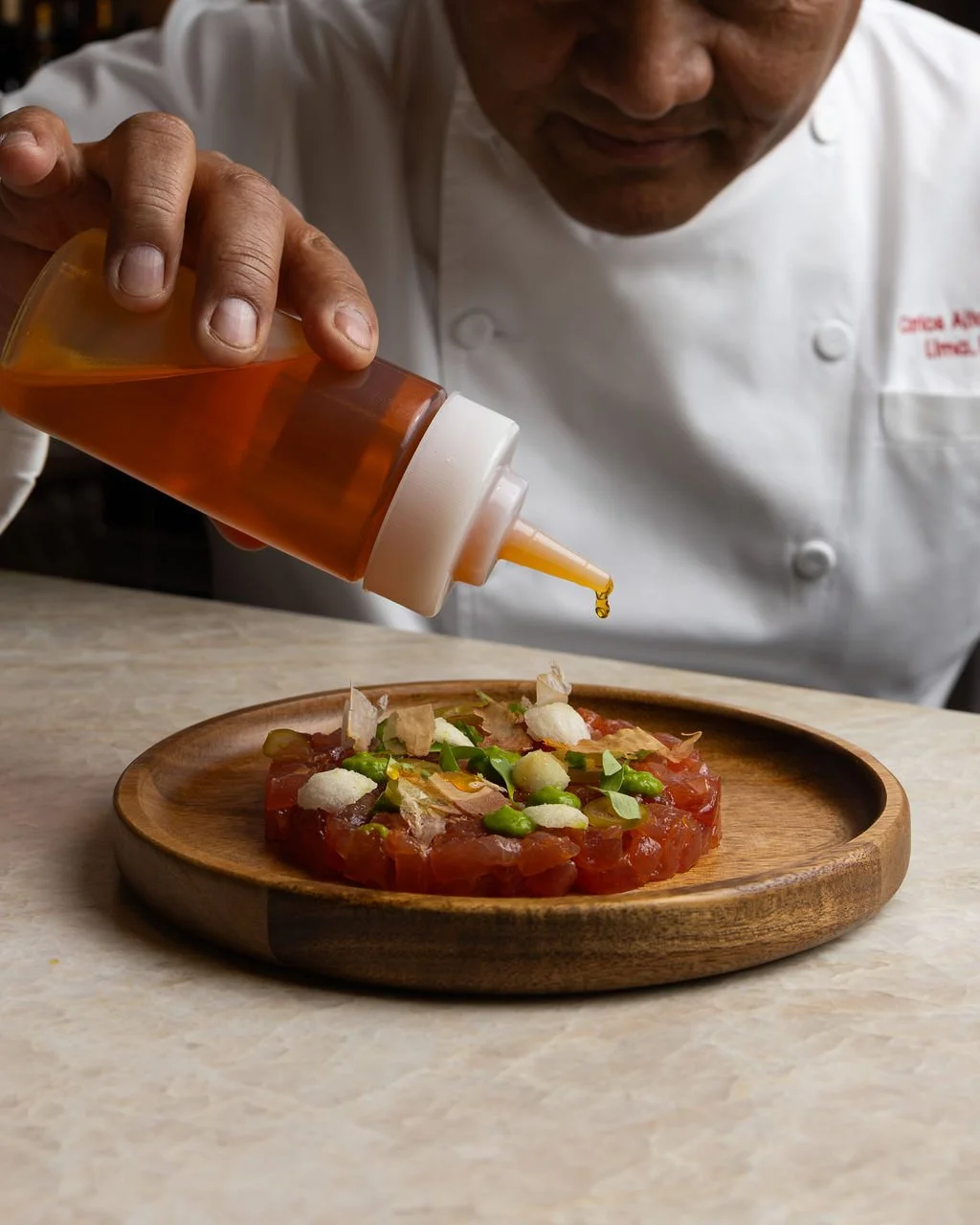 Chef pouring sauce over a raw fish tartare garnished with green onions and white onion flakes on a wooden plate.