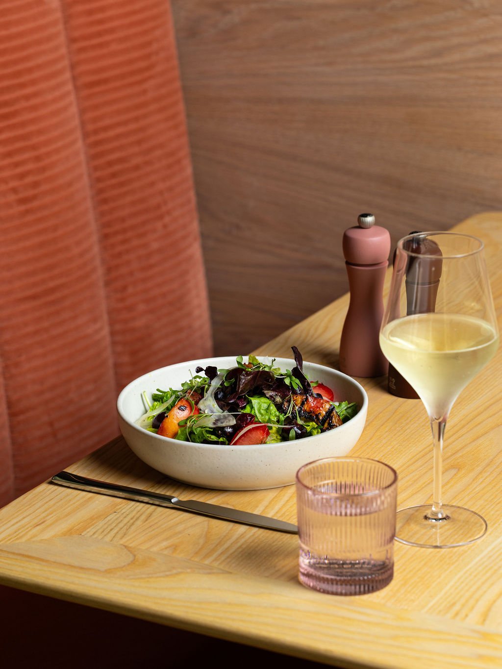 A wooden table with a bowl of mixed salad, a glass of white wine, a pink-tinted water glass, and pink salt and pepper shakers in the background.