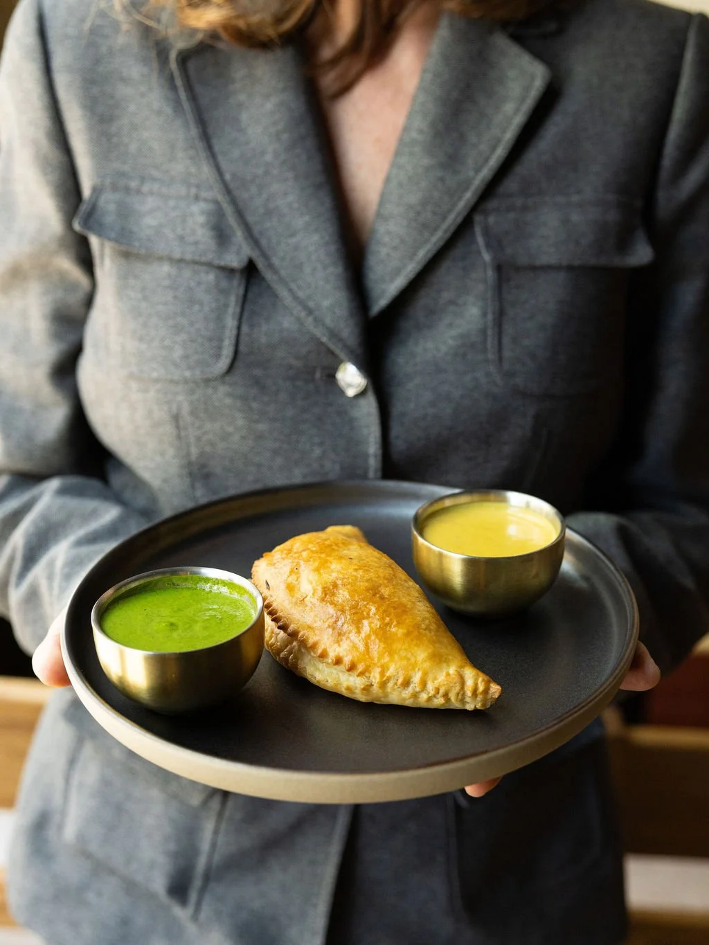 Person in gray blazer holding a black tray with a fried pastry and two small bowls of green and yellow sauces.