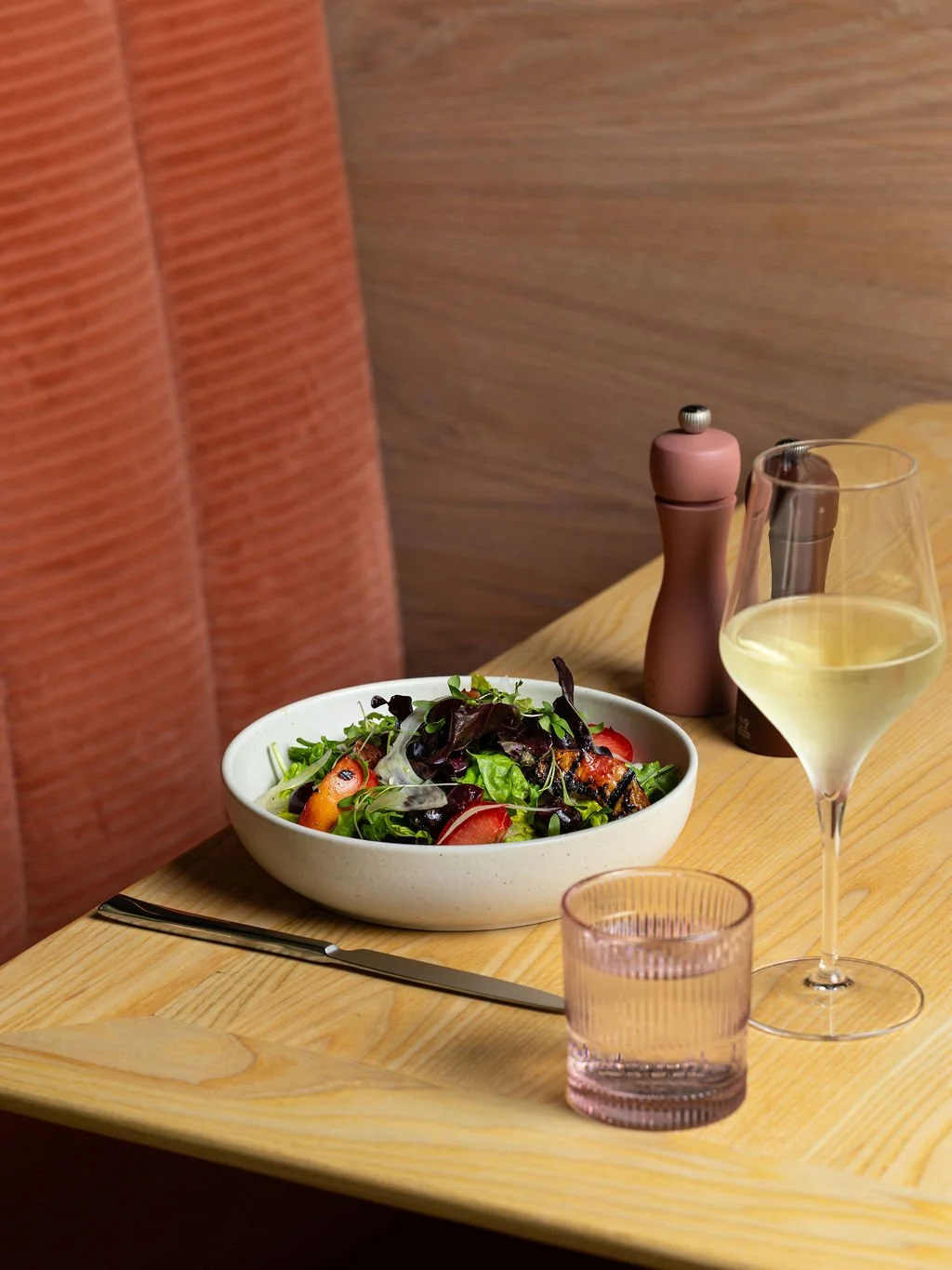 A wooden table with a bowl of mixed green salad, a glass of white wine, a pink salt shaker, and a pink-tinted glass of water, against a background of red and brown walls.