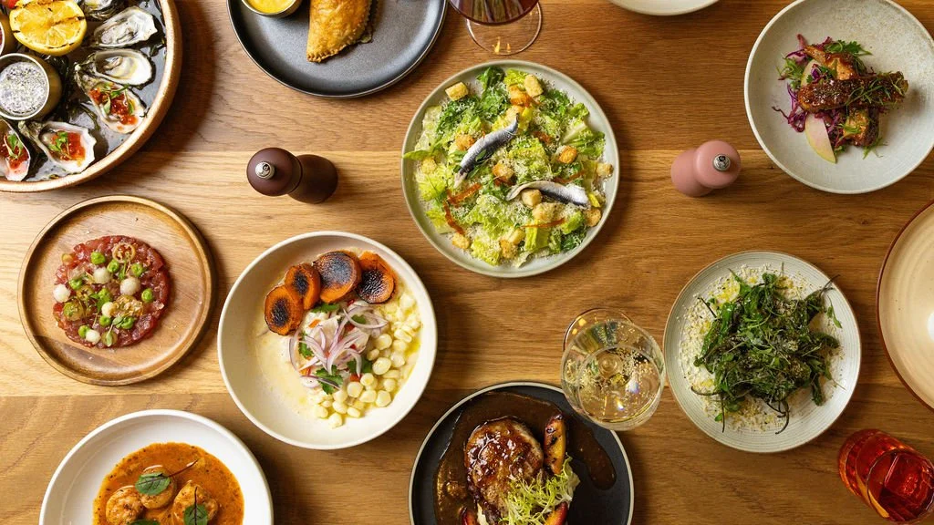 A variety of upscale dishes including oysters, a salad with anchovies, a steak, a dish with roasted vegetables, and small plates with meats and greens, set on a wooden table.