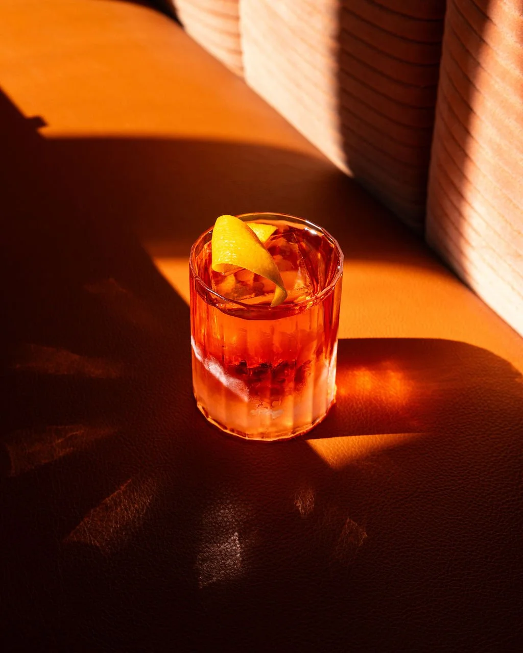 A glass of a pink-colored cocktail with ice cubes and a lemon twist garnish, illuminated by warm sunlight, on a textured surface.