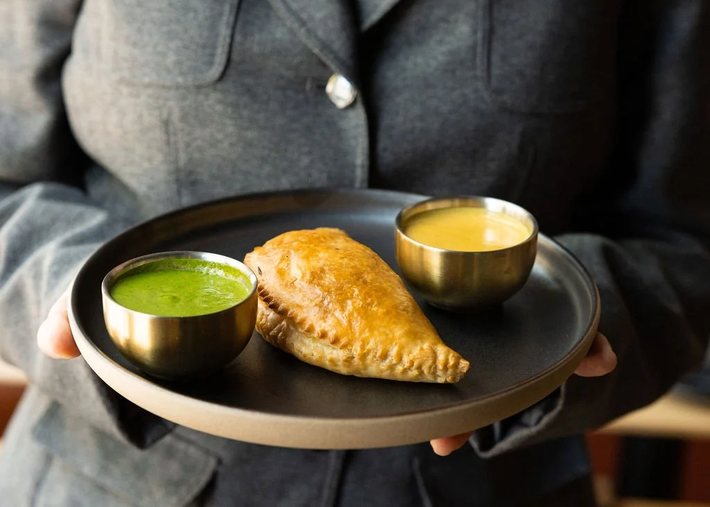 A person in a gray jacket holding a black plate with an samosa and two bowls of sauces, one green and one yellow.