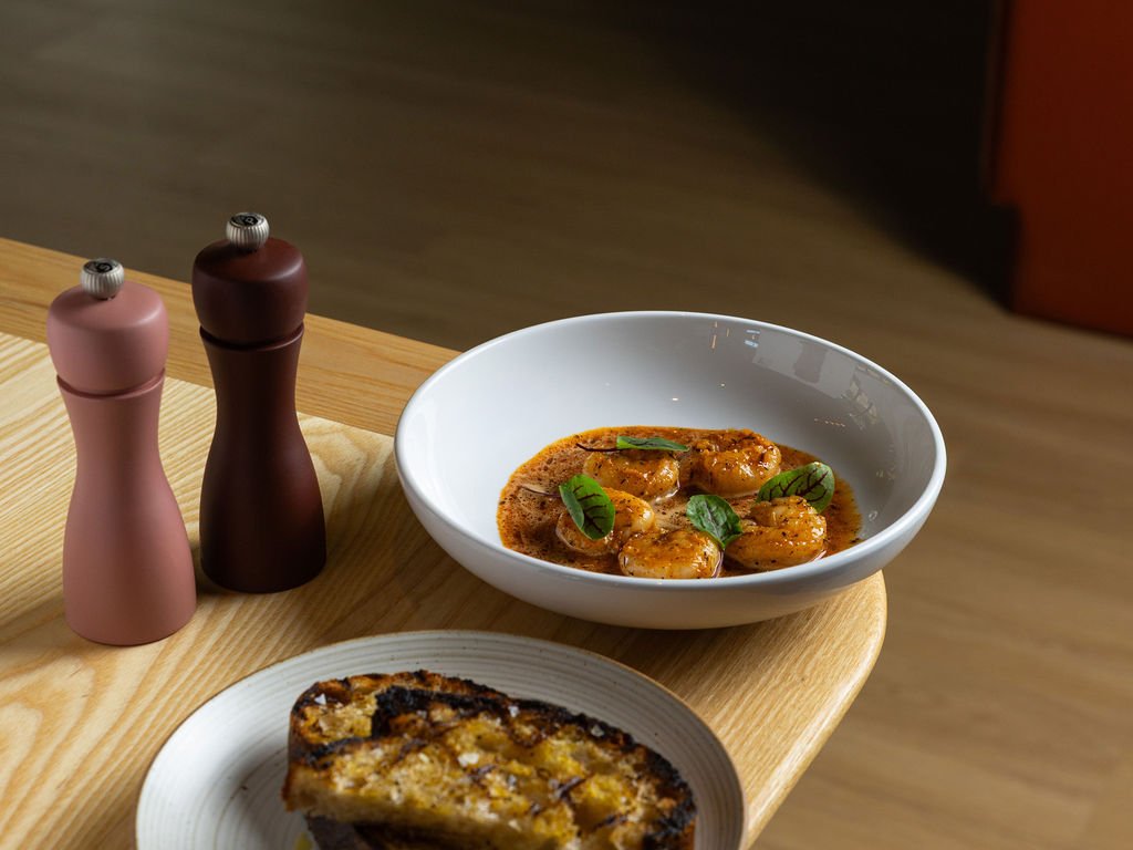 A bowl of shrimp in sauce garnished with basil leaves, a plate with a slice of quiche, and salt and pepper shakers on a wooden table.