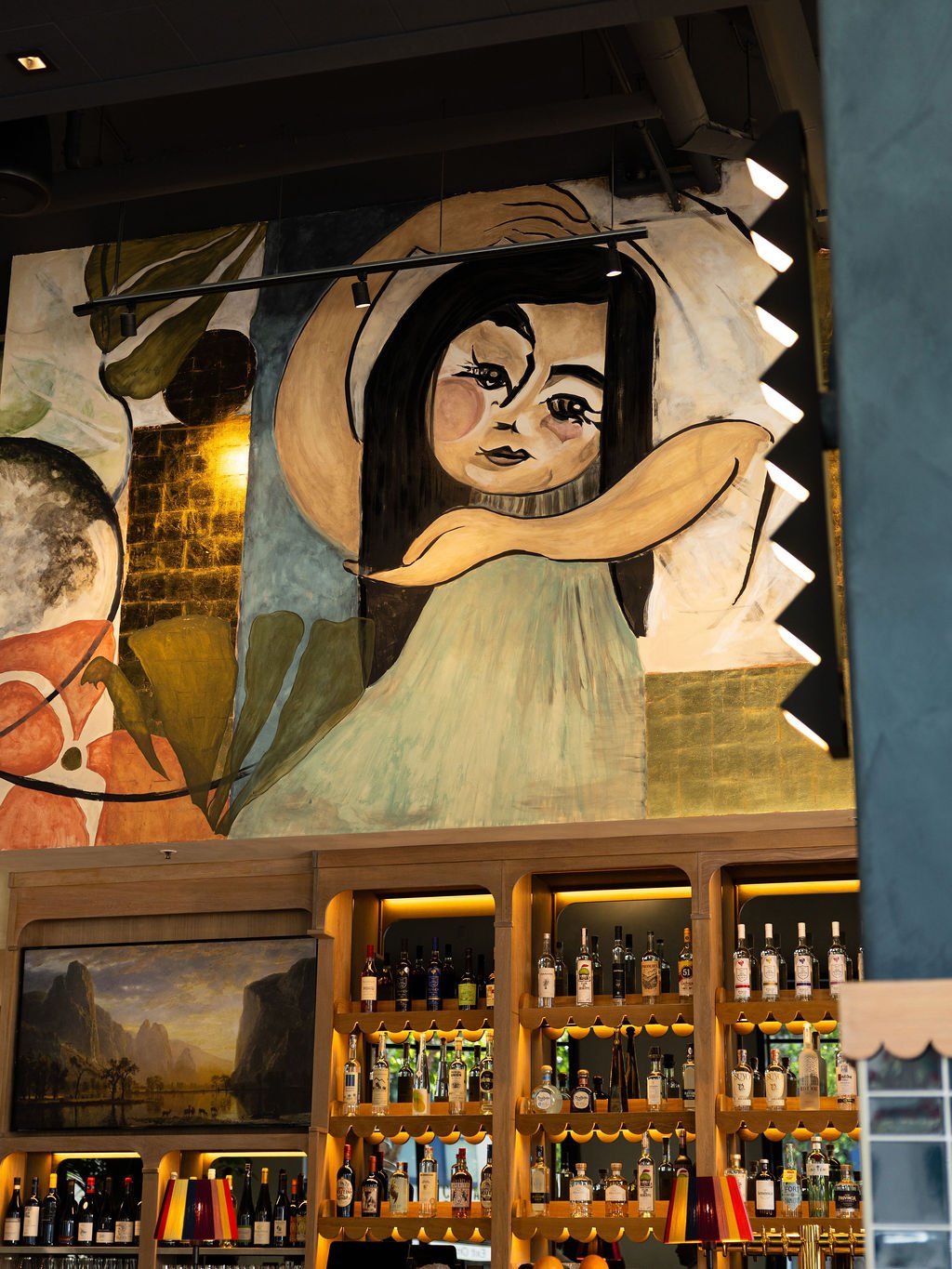 A large mural of a girl with long black hair holding her arm above her head, painted in a stylized, abstract manner, is visible on a wall above a bar with bottles of alcohol. The mural features warm, muted colors and is illuminated by nearby lights.