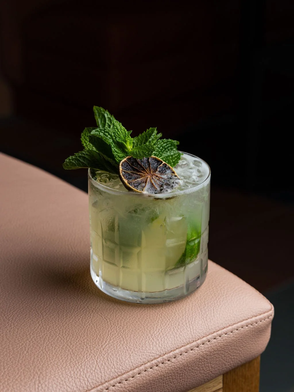 A glass of mojito cocktail with mint leaves and a charred lime wedge, placed on a pink upholstered surface.