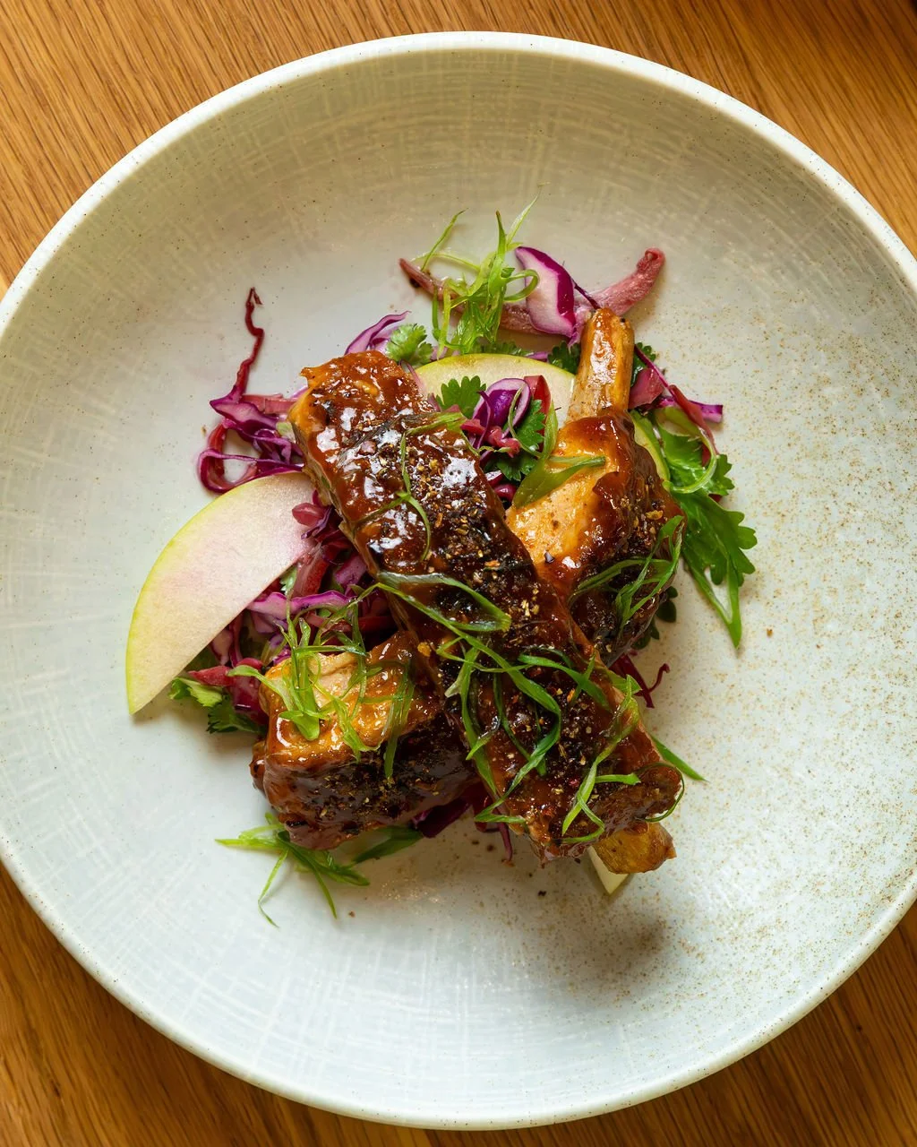A plated dish with glazed ribs on a bed of mixed greens, sliced apples, and garnished with herbs, on a white ceramic plate.