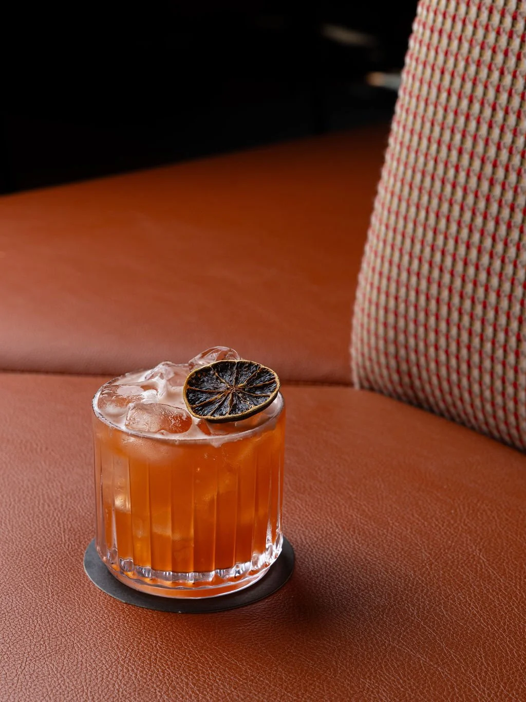 A glass of orange-colored cocktail with ice and a charred lemon slice on top, sitting on a black coaster on a brown leather surface.