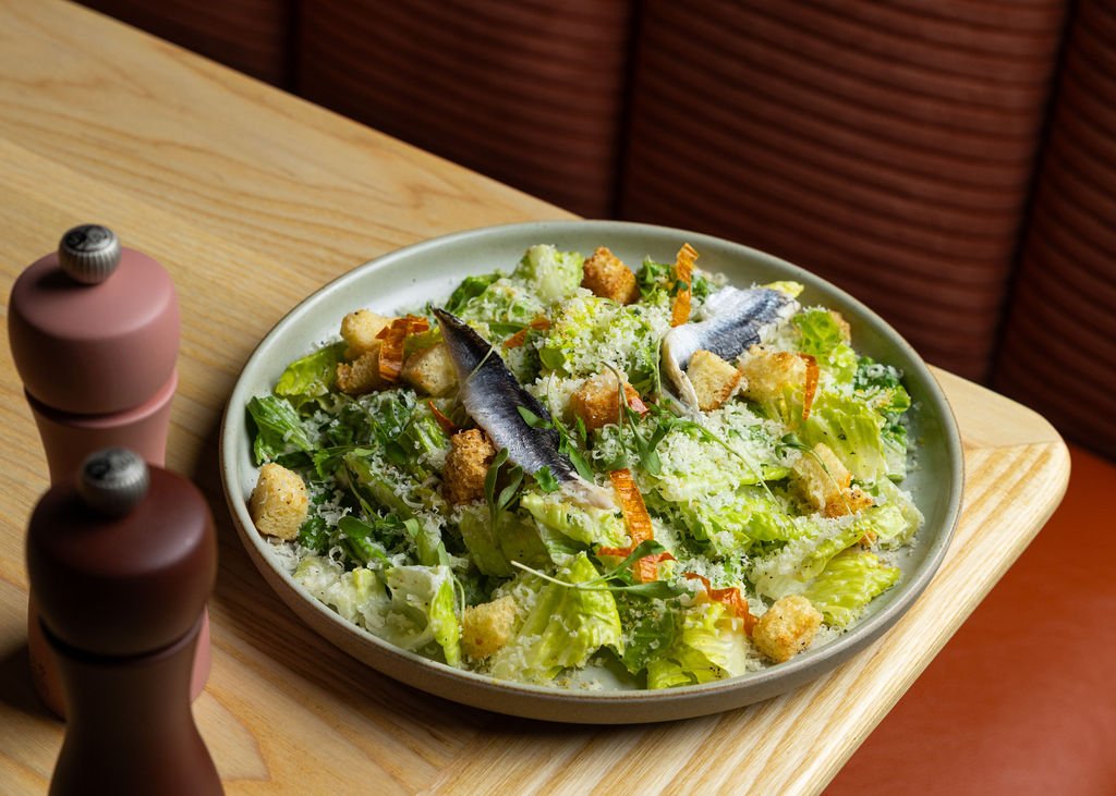 A Caesar salad with romaine lettuce, anchovies, croutons, grated cheese, and shredded carrots on a white plate at a wooden table.