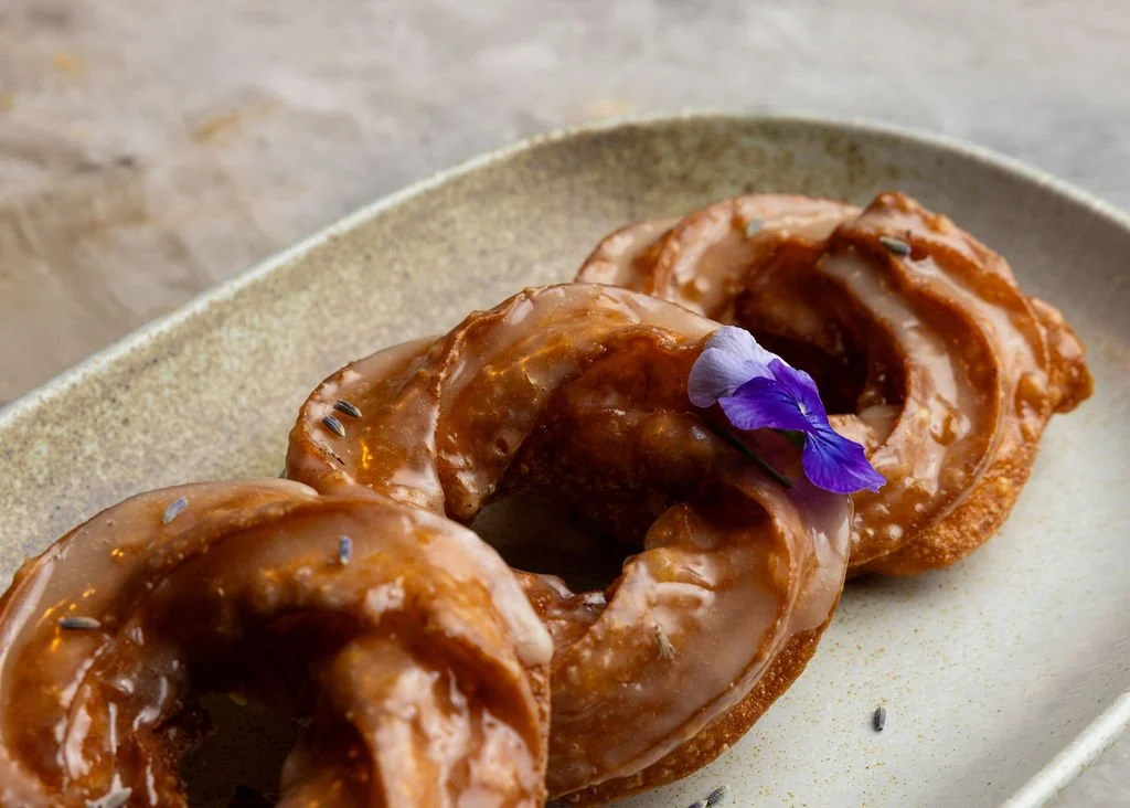 Three glazed donuts with a purple flower garnish on a beige plate