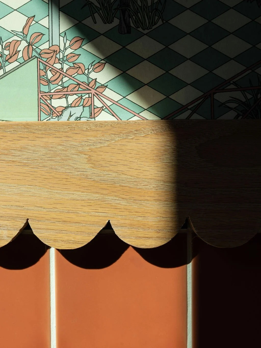 A wooden surface with scalloped edges partially blocking a colorful tiled floor with plants and a cat art illustration, casting shadows through a window.